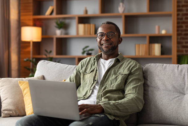 Feliz homem afro-americano de meia idade em óculos tem chamada de vídeo no laptop, surfando na internet no interior da sala de estar. Reunião remota, nova normal com gadget em casa, blog moderno, trabalho e webinar - Foto, Imagem