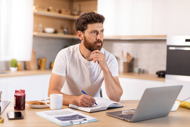 Pensive mature caucasian male with beard in white t-shirt thinks, works with documents, laptop in kitchen interior. Brainstorming, ideas for business, study and startup, device at home online lesson - Фото, изображение