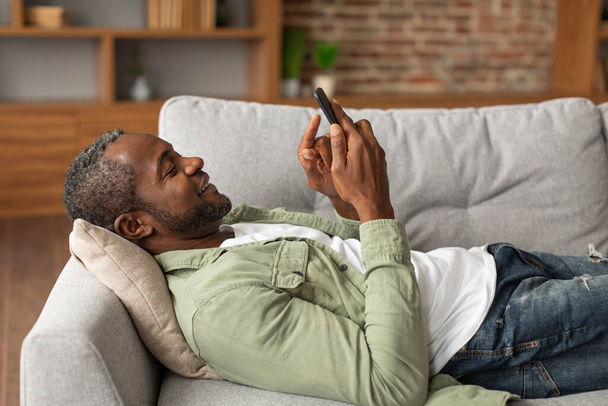 Sorrindo maduro afro-americano cara digitando no smartphone, escolhendo a ordem, conversando nas mídias sociais, encontra-se no sofá, desfrutar de tempo livre no interior da sala de estar. Descanse, relaxe com o aplicativo, assista a vídeos em casa - Foto, Imagem