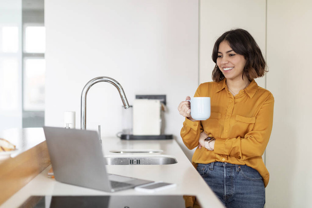 Mutfakta dizüstü bilgisayar kullanan ve kahve içen gülümseyen genç bir Arap kadın, bilgisayardan video izleyen ve evinde kafein içen mutlu bir Orta Doğulu kadın, - Fotoğraf, Görsel