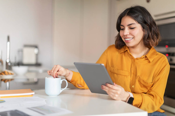 Jovem mulher árabe feliz relaxando com tablet digital na cozinha e beber caféEm casa, sorrindo Médio Oriente Feminino Navegando Internet no Gadget moderno e desfrutando de bebida quente, espaço livre - Foto, Imagem
