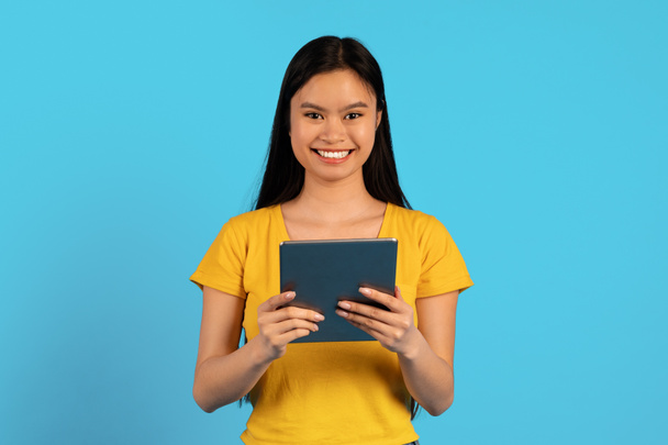 Sarı tişörtlü neşeli Çinli kız öğrenci tablete yazı yazıyor, mavi arka planda izole edilmiş video izliyor, stüdyo. Bağımsızlık, çalışma ve öğrenim aracı ve uygulaması, reklam ve teklif, çevrimiçi satış - Fotoğraf, Görsel