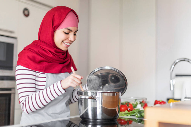 Joven mujer árabe alegre que usa hijab, delantal y ropa casual cómoda ama de casa cocinando en la cocina eléctrica en casa, mezclando comida en olla y sonriendo, espacio para copiar. Cocina, concepto de estilo de vida doméstico - Foto, Imagen