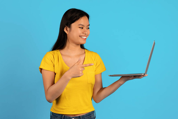Реклама і пропозиція, величезний розпродаж. Щаслива молода азіатська студентка в жовтій футболці вказує пальцем на екран комп'ютера ізольовано на синьому фоні. Рекомендації, поради професіонала, пристрій для роботи, навчання
 - Фото, зображення