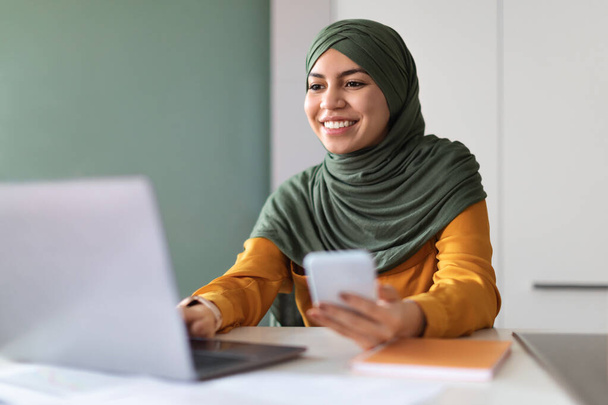 Retrato de una mujer árabe joven en Hijab usando un teléfono inteligente y un ordenador portátil mientras trabaja en casa, una mujer musulmana milenaria con pañuelo en la cabeza sentada en el escritorio y escribiendo en la computadora, disfrutando de un trabajo en línea - Foto, Imagen