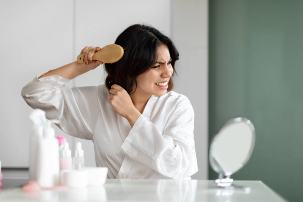 Hair detangling concept. Angry middle eastern young lady in silk robe brushing dry hair with wooden hairbrush, suffer from pain during beauty routine, sitting at beauty table at home, look at mirror - Foto, afbeelding