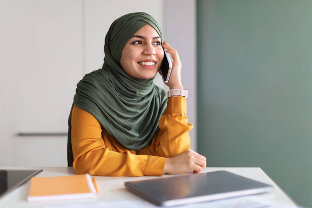 Portrait Of Smiling Muslim Woman In Hijab Talking On Mobile Phone In Office, Happy Young Islamic Lady In Headscarf Enjoying Pleasant Conversation On Cellphone And Looking Away, Copy Space - 写真・画像