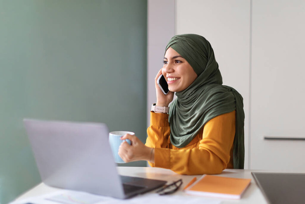 Smiling Muslim Woman Talking On Cellphone And Drinking Coffee At Workplace In Office, Young Islamic Lady In Hijab Sitting At Desk With Laptop And Having Mobile Conversation, Enjoying Break At Work - Photo, Image