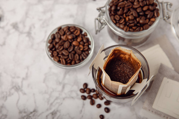 自宅でコーヒーを作る傾向があります。茶色の背景にコーヒー豆とガラスに新鮮な飲み物の袋をドロップします。カップで醸造するためのグラウンドコーヒー。ペーパータイプフィルターを使用したドリップコーヒー. - 写真・画像