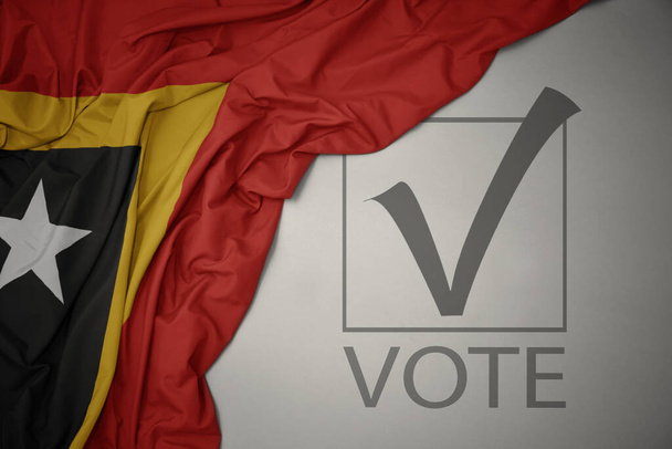 ondeando colorida bandera nacional de Timor Oriental sobre un fondo gris con voto de texto. concepto de elección. Ilustración 3D - Foto, imagen