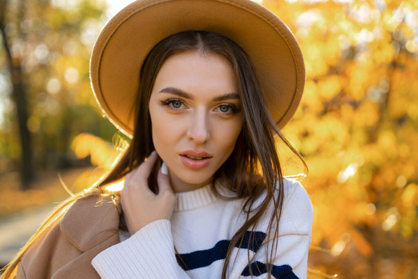 portret van aantrekkelijke stijlvolle lachende vrouw met lang haar wandelen in het park, gekleed in warme bruine jas herfst trendy mode, straat stijl dragen hoed. - Foto, afbeelding