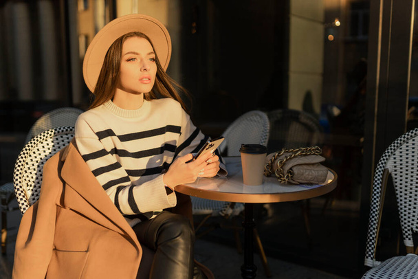 Menina morena alegre atraente em casaco de trincheira com café para ir feliz usando celular ao ar livre.  - Foto, Imagem