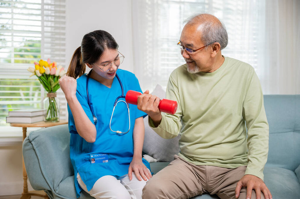 Young nurse physiotherapist helping old man in lifting dumbell at home, Asia nurse support elderly retirement sitting on sofa using red dumbbell workout exercise physical recovery, Health care concept - Photo, Image