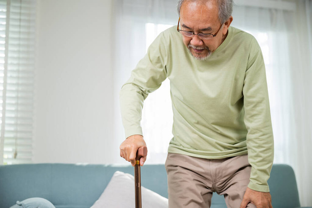 Asian Old man with eyeglasses typing to stand up from sofa with walking cane, Elderly suffering from knee pain ache holding handle of cane, senior disabled man holding walking stick at home - Photo, Image