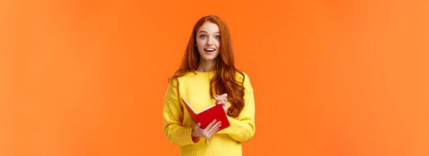 Geïnspireerd en vrolijk, dromerig gefascineerd roodharig meisje dat interessante lezingen opschrijft, glimlacht en staart met bewondering of enthousiasme, invulnotitieboekje, staande oranje achtergrond. - Foto, afbeelding