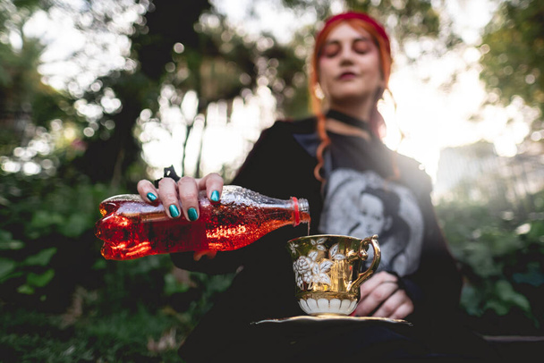 Fiatal, vörös hajú lány vörös sállal és sötét ruhával, aki egy régi évszázados teáscsészében és csészealjban szolgál fel vörös italt egy üvegből a parkban. - Fotó, kép