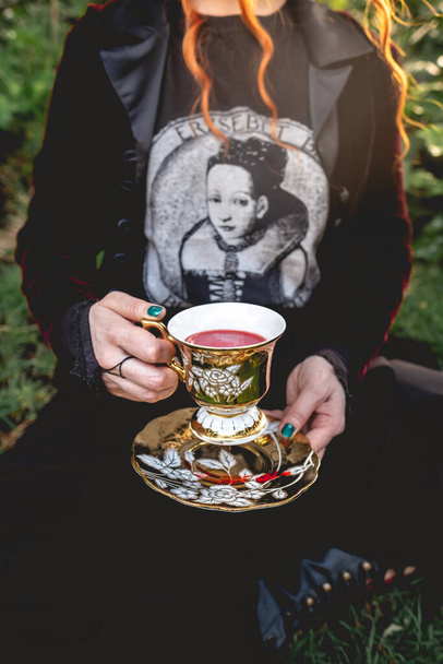 Rothaariges Mädchen mit dunkler Kleidung (und ohne Kopf), das ein rotes Getränk ("Blut") in einer alten Teetasse und Untertasse im Park trinkt - Foto, Bild