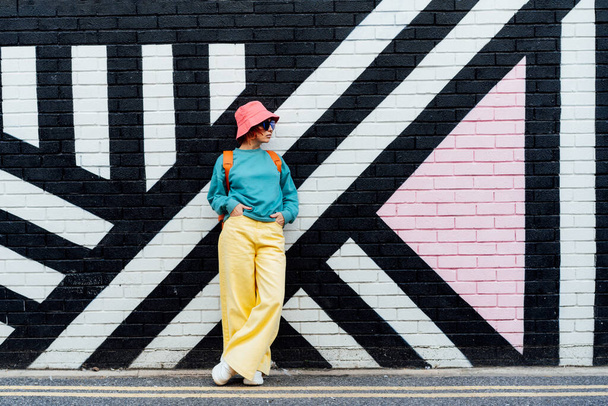 Hipster fashion young woman in bright clothes, sun glasses and bucket hat posing on the painted brick wall background. Urban city street fashion. Selective focus. Copy space. - Fotó, kép