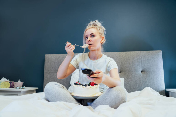 Surprised, stressed, shocked woman eating cake and holding a remote control while watching TV in bed. Scary horror movie or terrible news, scare in her eyes Overeating, Jamming of negative emotions. - Foto, Imagen