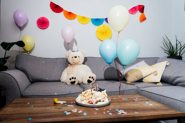 Messy table in living room after party. Broken birthday cake, confetti, balloons and decoration. Big teddy bear sitting on the sofa. Selective focus - Photo, image