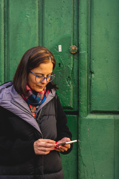 Женщина улыбается, когда она взаимодействует со своим смартфоном рядом с зеленой деревянной дверью - Фото, изображение