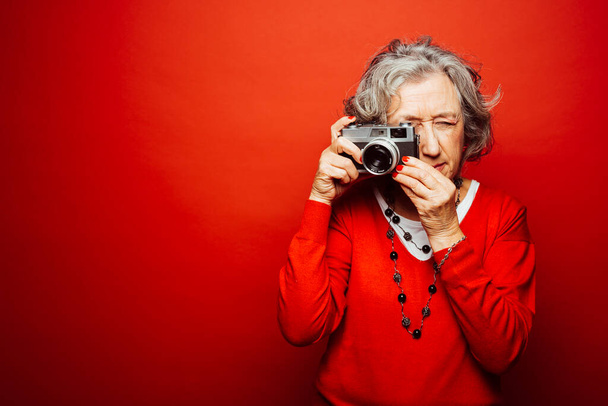 Старшая женщина в красной одежде, фотографирующая со старой кинокамерой, на красном фоне - Фото, изображение