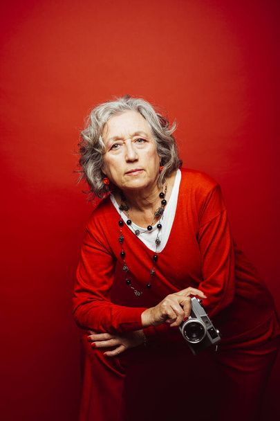 Ηλικιωμένη γυναίκα που φοράει κόκκινα ρούχα, βγάζει φωτογραφίες με μια παλιά φωτογραφική μηχανή, πάνω από ένα κόκκινο φόντο - Φωτογραφία, εικόνα