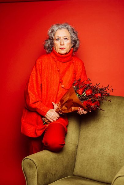 Старшая женщина в красной одежде и сидит на зеленом кресле с букетом красных роз, на красном фоне - Фото, изображение