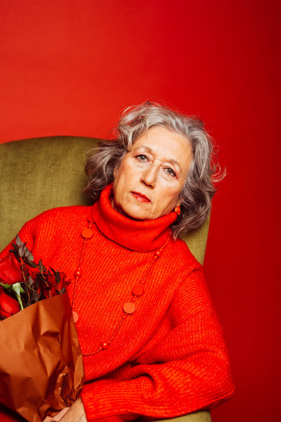 Старшая женщина в красной одежде и сидит на зеленом кресле с букетом красных роз, на красном фоне - Фото, изображение