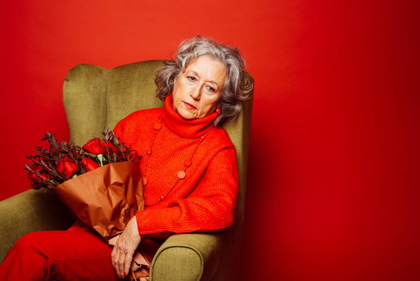 Старша жінка в червоному одязі і сидить на зеленому кріслі з букетом червоних троянд, на червоному тлі
 - Фото, зображення