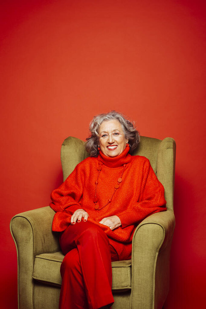 Starsza kobieta ubrana w czerwone ubrania i siedząca na zielonym fotelu na czerwonym tle - Zdjęcie, obraz