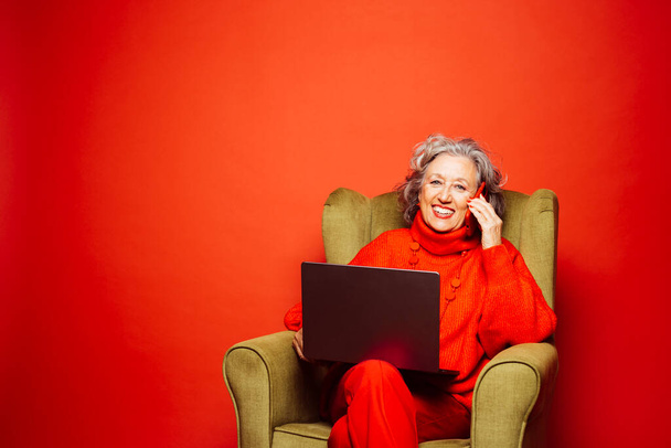 Kıdemli kadın, kırmızı elbiseler giyip yeşil koltukta oturuyor, kırmızı arka planda dizüstü bilgisayar ve telefon kullanıyor. - Fotoğraf, Görsel