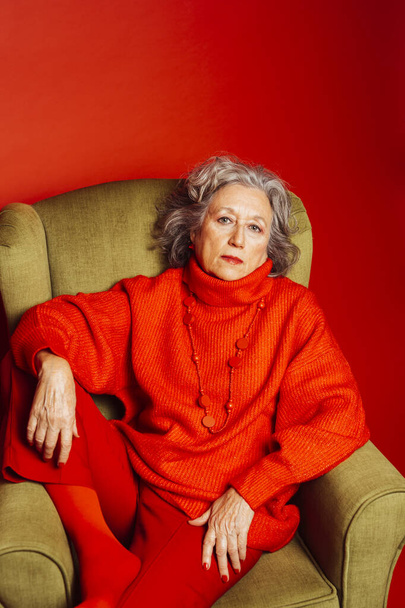 Senior woman wearing red clothes and sitting on a green armchair over a red background - Фото, изображение