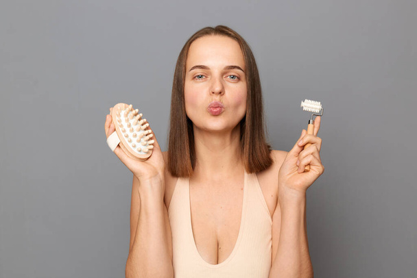 Funny positive woman showing dry body brush and massage roller, wants to have smooth clean skin, smiles gladfully enjoys part of morning health and energy routine, poses against gray background. - Foto, Imagen