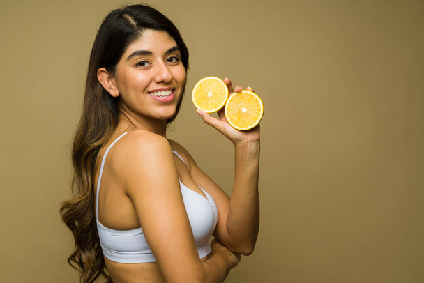 Perfil de uma jovem alegre usando um sutiã branco tomando vitamina c e comendo frutas de laranja para uma boa saúde e pele clara  - Foto, Imagem