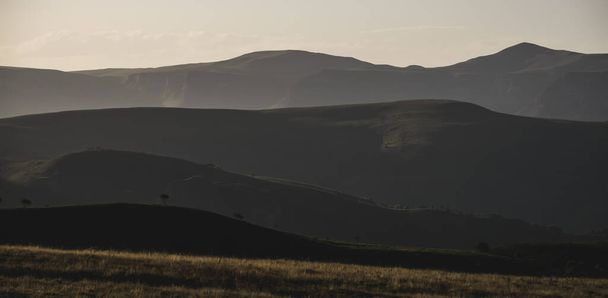 Панорама горных хребтов с холмами и растительностью на них, при вечернем освещении заката - Фото, изображение