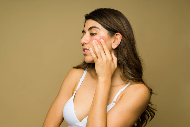 Mujer hermosa hispana poniéndose una mascarilla facial para hidratar su piel sana contra un fondo de estudio - Foto, Imagen