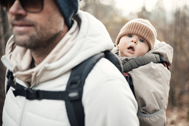 Sportovní otec nesoucí svého malého syna v zimní kombinéze a čepici v batohu nosič turistiky v podzimním lese - Fotografie, Obrázek