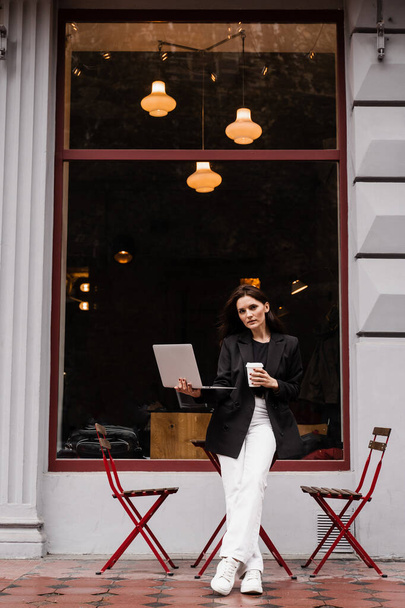 Wesoła dziewczyna z laptopem stojąca niedaleko kawiarni. Młoda kobieta trzyma laptopa i uśmiecha się w kawiarni - Zdjęcie, obraz