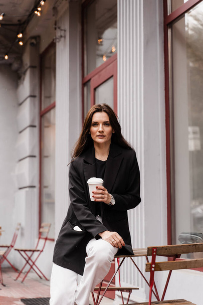 Уверенная молодая деловая женщина с чашкой кофе позирует перед кафе на террасе. Веселая девушка носится случайный бизнес стиль позирует возле кафе - Фото, изображение