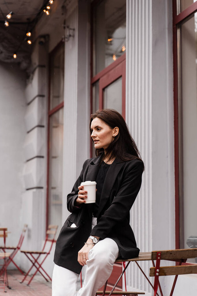 Důvěryhodná mladá podnikatelka s šálkem kávy pózuje mimo kavárnu na terase. Veselá dívka je nosit ležérní obchodní styl je pózování v blízkosti kavárny - Fotografie, Obrázek