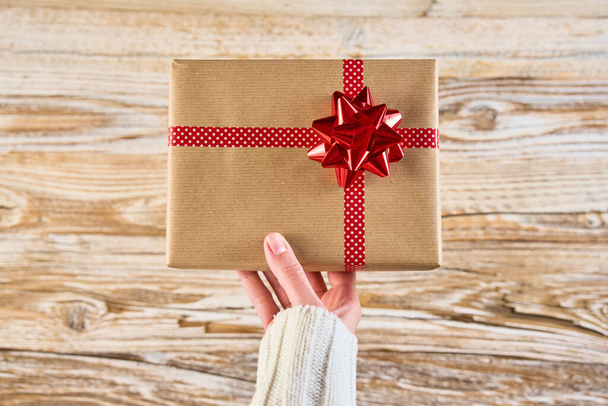 Vrouw houdt geschenkdoos verpakt in ambachtelijk papier met feestelijk lint tegen houten achtergrond. Vrouwelijke handen houden cadeau voor kerstvakantie, boven het zicht - Foto, afbeelding