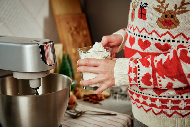 Vrouw in feestelijke kersttrui die thuis deeg maakt voor koekjes. Vrouwelijke handen gebruiken een elektrische mixer. Moderne keukenapparatuur - Foto, afbeelding