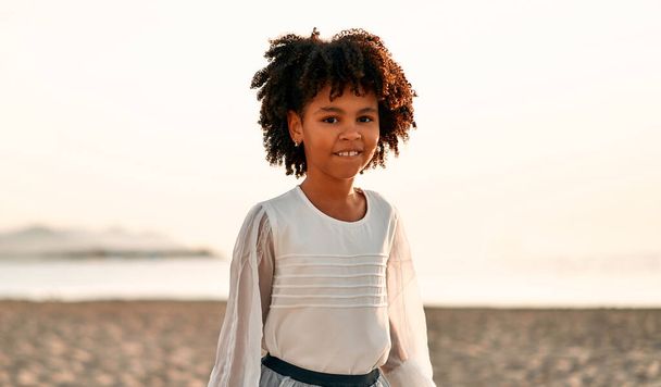 Cute African American child girl in a white dress with afro curls standing on a sandy beach by the sea in sunny cloudless weather. - Foto, immagini