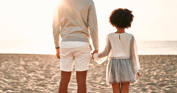 Afrika kökenli Amerikalı bir aile deniz kenarındaki kumlu sahilde eğleniyor. Beyaz elbiseli tatlı kızıyla genç bir baba el ele tutuşan deniz manzarasına bakıyor.. - Fotoğraf, Görsel