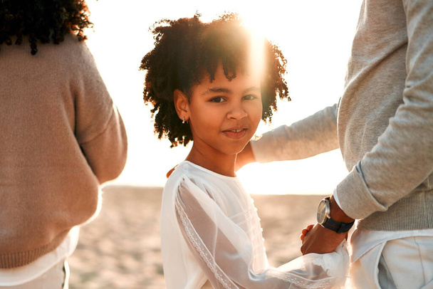 Famille afro-américaine avec une jolie fille bouclée dans une robe blanche tenant la main marchant le long de la plage de sable près de la mer à l'aube. - Photo, image