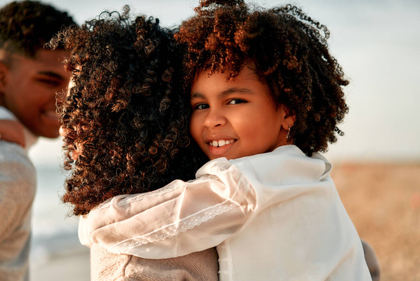 African American woman mom with her cute daughter with afro curls standing hugging on the seashore at dawn on their weekend. - Zdjęcie, obraz