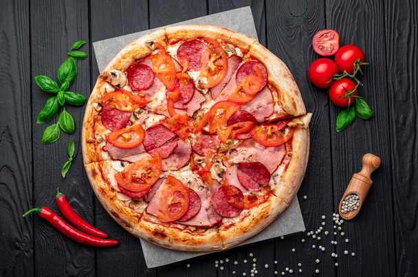 Italienische Pizza mit Schinken, Wurst, Pilzen, Tomaten und Zwiebeln. Italienische Pizza auf dunklem Hintergrund. Foto von Pizza für Speisekarte - Foto, Bild