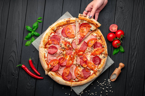 Pizza italiana con prosciutto, salsiccia, funghi, pomodori e cipolle. Pizza italiana su sfondo scuro. Foto di pizza per menu - Foto, immagini