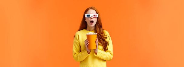 Shocked and tensed redhead woman seeing intense fight on screen as watching movie at cinema, wear 3d glasses, holding popcron, frowning and gasping concerned, orange background. - Foto, imagen